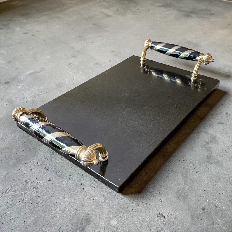 May include: A rectangular black serving tray with gold-coloured handles. The handles have a black and silver striped design. The tray is made of a solid, dark material, and the handles are ornate.