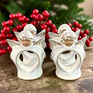 May include: Two ceramic napkin rings shaped like angels with iridescent white glaze and gold-toned accents on the wings and hair. Each angel features a circular opening in the center for holding a napkin, with a smooth, glossy finish throughout.