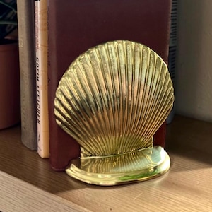 May include: A gold-colored seashell-shaped bookend. The bookend is made of metal and has a detailed, textured surface. It is positioned between two books on a wooden shelf, with a potted plant visible in the background.