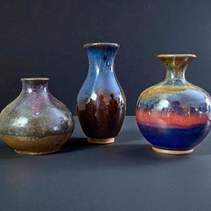 May include: Three ceramic vases with a glossy finish. The vases have a variety of colors, including blue, brown, and red. The vases are of different sizes and shapes, with a rounded body and a narrow neck. They are displayed on a dark surface.