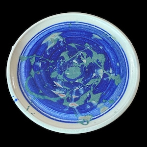May include: A round ceramic plate with a white rim and a blue and green abstract design. The center of the plate features a swirling pattern of blue and green, with hints of red and white. The plate is set against a black background.