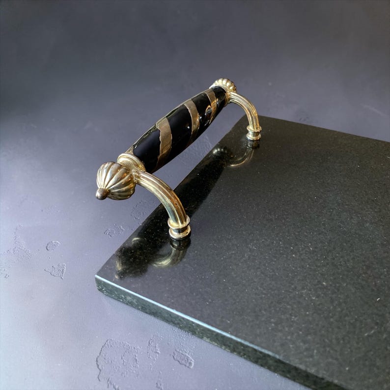 May include: A black serving tray with a decorative handle. The handle is black with gold accents and gold-coloured hardware. The tray is rectangular and made of a dark, possibly stone-like material.
