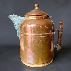 May include: A vintage copper tea kettle with a wooden handle and a decorative spout. The kettle has a round body and a lid with a small knob. The spout is a light blue color. The handle is made of dark wood and brass.