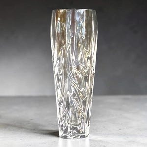 May include: A tall, clear glass vase with a textured, swirling pattern. The vase is cylindrical with a slightly flared top and sits on a gray surface. The design creates a rippled effect, adding visual interest to the decorative piece.