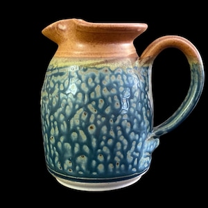 May include: A handmade ceramic pitcher with a textured, mottled blue glaze. The rim and handle are a warm brown, contrasting with the blue body. The pitcher has a spout for easy pouring and a sturdy handle.