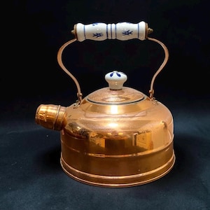 May include: A polished copper tea kettle featuring a rounded body and a short spout. The kettle includes a white ceramic handle and a matching lid knob, both decorated with small blue painted patterns. The metal surface reflects light.