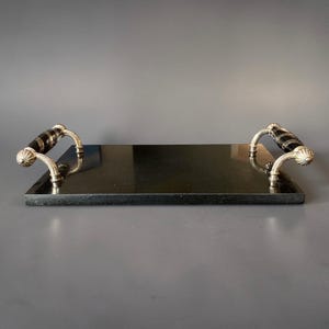 May include: A rectangular black serving tray with silver-coloured handles. The handles have a decorative design with black accents. The tray is made of a dark, possibly stone-like material.