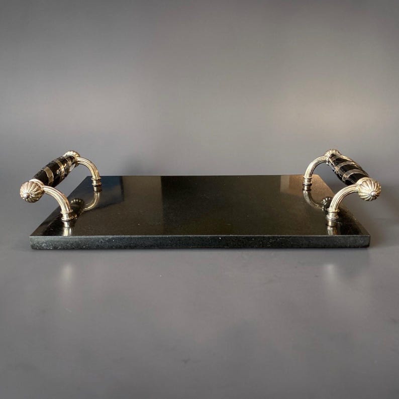 May include: A rectangular black serving tray with silver-coloured handles. The handles have a decorative design with black accents. The tray is made of a dark, possibly stone-like material.