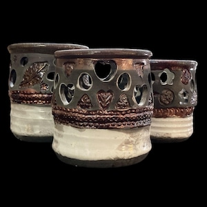 May include: Three ceramic candle holders with a dark gray finish, featuring heart and leaf cutouts. The holders have a white base and a textured, dark brown band. The rims are also dark brown, creating a rustic aesthetic.