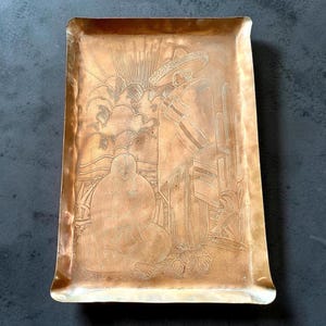 May include: Rectangular copper-colored tray with a detailed etched design. The design depicts a figure wearing a sombrero, a cactus, and a seated figure. The tray has a slightly raised edge and rounded corners.
