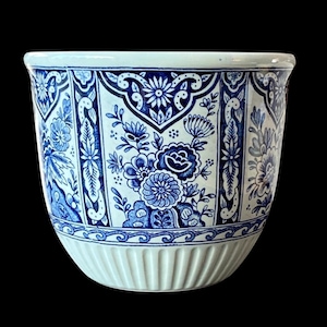 May include: A white ceramic planter with a blue floral design. The planter features a repeating pattern of flowers and leaves in shades of blue. The bottom of the planter has a ribbed texture.