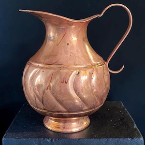 May include: A copper pitcher featuring a rounded body with a subtle swirling embossed pattern, a flared rim, and a curved handle. The metal surface displays a polished finish with natural variations in color and texture throughout the exterior.