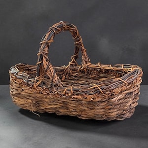 May include: A rectangular woven basket crafted from dark brown natural fibers. The basket features a sturdy, arched handle made of twisted vines and a thick, braided rim. The base is constructed from tightly woven, textured organic material.