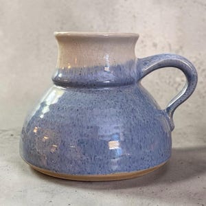 May include: A handmade ceramic mug with a blue and tan glaze. The mug has a rounded body, a handle, and a flared rim. The blue glaze has a speckled appearance, and the rim is tan. The mug is suitable for beverages.