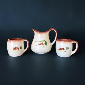 May include: A ceramic creamer pitcher and two matching mugs featuring a cream-colored base with a brown gradient rim. Each piece is decorated with a small hand-painted scene of a red barn, green trees, and rolling hills on the front surface.