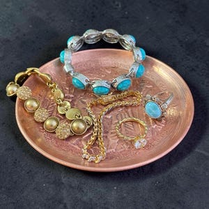 May include: A round copper-colored tray holding a collection of jewelry including a silver-toned bracelet with turquoise-colored stones, a gold-toned bracelet with textured circular links, a thin gold-toned chain, and two rings with blue stones.