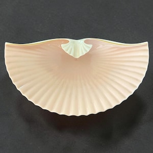 May include: A pale pink, fan-shaped decorative dish with a ribbed texture. The dish has a light green accent at the center and a thin gold trim along the top edge. The dish is likely made of ceramic or porcelain.