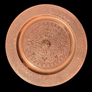 May include: A copper-colored plate with an embossed Aztec calendar design. The plate features intricate details of the calendar's symbols and patterns, with a circular border of additional designs. The plate is set against a black background.