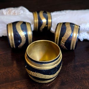 May include: A set of four cylindrical napkin rings featuring a dark, matte black finish contrasted with ornate, raised gold-toned bands. Each ring displays a central wavy, embossed pattern and smooth, metallic gold-toned edges.