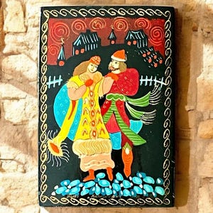 May include: A black wooden box with a painted scene of a couple in traditional clothing standing in front of a village. The woman is wearing a yellow and blue dress and the man is wearing a red and green outfit. The background is painted in shades of red, orange, and blue.