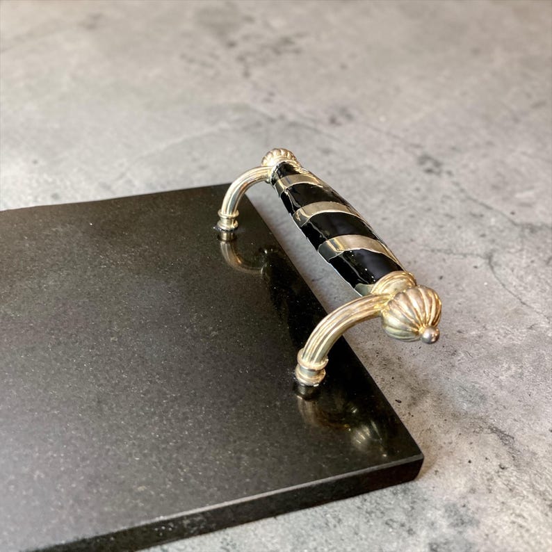 May include: A rectangular black serving board with a decorative handle. The handle is silver-toned with a black and silver striped design. The board is made of a dark, possibly stone-like material.