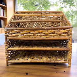 May include: A handcrafted, multi-tiered desk organizer made of woven bamboo. The organizer has a house-like shape with a sloped roof and three open shelves. The natural, light brown color of the bamboo gives it a rustic, yet elegant appearance.