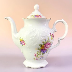 May include: White porcelain teapot with a lid, spout, and handle. The teapot is decorated with floral designs in pink, purple, yellow, and blue. The teapot has a decorative embossed pattern.