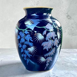 May include: A deep navy blue vase featuring a hand-painted design of cascading wisteria flowers in shades of light blue, lavender, and white. The vase has a smooth, glossy finish and a rounded body with a narrow neck and a thin, light-colored rim.