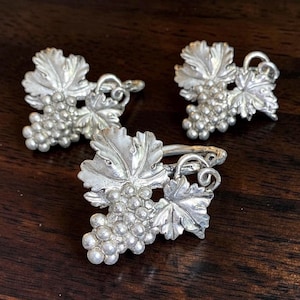 May include: Three silver-toned metal napkin rings, each featuring a detailed three-dimensional cluster of grapes and textured vine leaves. The metallic finish has a polished sheen, highlighting the intricate embossed patterns on the fruit and foliage.