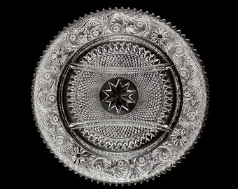 May include: Clear glass serving dish with a detailed floral and starburst pattern. The dish has a divided center section and a scalloped edge. The design includes flowers, leaves, and geometric shapes, creating an elegant and decorative piece.