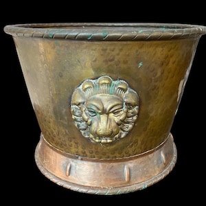 May include: A hammered brass planter with a lion's head emblem. The planter has a wide, textured body with a decorative rim and base. The lion's head is in the center of the planter, with a detailed mane and facial features.