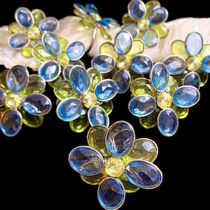 May include: A set of five flower-shaped napkin rings featuring faceted oval blue and lime green glass stones set in silver-toned metal frames. Each piece is arranged in a circular pattern with a central stone surrounded by six petals.