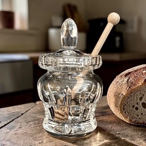 May include: A clear glass honey jar with a lid and a wooden honey dipper. The jar has a decorative, cut-glass design. A loaf of bread is on the right side of the jar. The jar is sitting on a wooden surface.