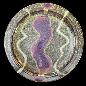 May include: A decorative ceramic plate with a dark brown rim and a speckled gold interior. A central purple design with red accents is flanked by two white wavy lines. The plate is round.