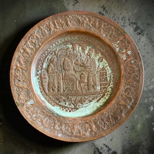 May include: A round, antique copper plate with an embossed design. The center features a detailed relief of figures, possibly a historical scene. The outer rim has a decorative floral pattern. The plate shows signs of age with a green patina.