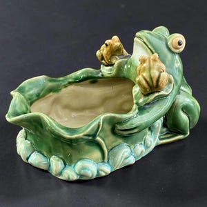 May include: A glazed ceramic planter shaped like a green lily pad with a large frog sitting on the edge and two smaller frogs perched on the rim. The interior is a smooth cream color, while the exterior features shades of green and blue.