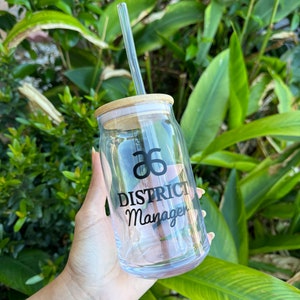 Puede incluir: Un vaso de vidrio transparente con tapa de bambú y pajita transparente. El vaso presenta el texto "District Manager" en escritura negra. El fondo es follaje verde.