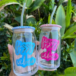 May include: Two clear glass tumblers with lids and glass straws. One tumbler has a blue design that says "fizzy vibes", and the other has a pink design that says "fizzy vibes". The lids are decorated with rhinestones.