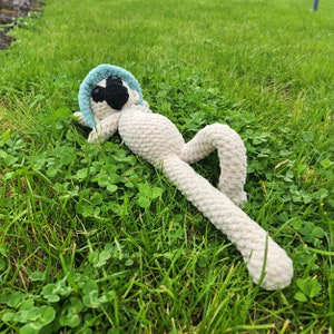 Peut inclure: Un animal en peluche crocheté ressemblant à un lémurien, allongé sur le dos dans un champ herbeux. Le lémurien est blanc avec un chapeau bleu et des yeux noirs.