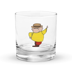 Op de afbeelding: Een helder tumblerglas met een cartoonillustratie van een man in een geel shirt, rode broek en een bruine hoed. Het personage gebaart met één arm omhoog. Het glas is ontworpen voor dranken.