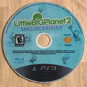 May include: A blue and green PlayStation 3 game disc for LittleBigPlanet 2 Special Edition. The disc has a blue background with white text and a green logo. The disc is surrounded by a white border.