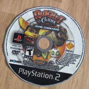 May include: Circular rug designed to resemble a PlayStation 2 game disc. The rug features the game title "Ratchet & Clank: Going Commando" with character illustrations. The design includes the PlayStation 2 logo and game details.