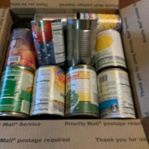 May include: A cardboard box filled with various canned goods. The cans are labeled with different food items, including peaches, corn, and green beans. The box has a label that says "Priority Mail" and "postage required".