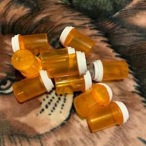 May include: A collection of ten empty, translucent orange plastic pill bottles with white snap-on caps. The cylindrical containers are arranged in a cluster, showing their semi-transparent orange bodies and opaque white lids from various angles.