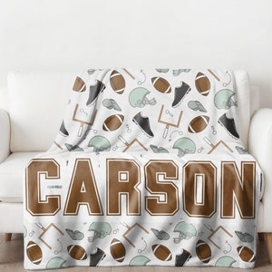Op de afbeelding: Een witte deken met een herhaald patroon van American footballs, helmen, schoenplaatjes en doelpalen. De naam "CARSON" staat in grote bruine letters onderaan de deken.
