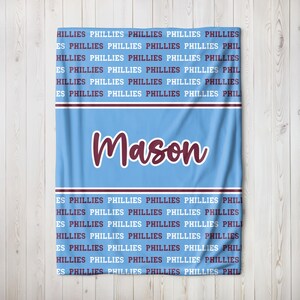 May include: Light blue blanket with the name "Mason" in maroon script. The blanket is patterned with the word "PHILLIES" in maroon. A maroon and white stripe design is at the top and bottom of the blanket.