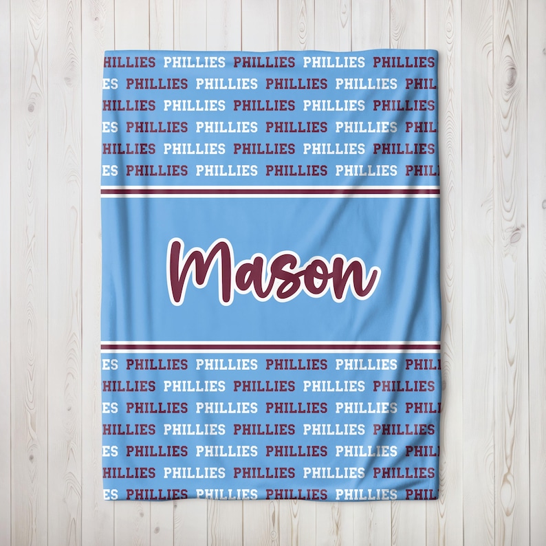 May include: Light blue blanket with the name "Mason" in maroon script. The blanket is patterned with the word "PHILLIES" in maroon. A maroon and white stripe design is at the top and bottom of the blanket.