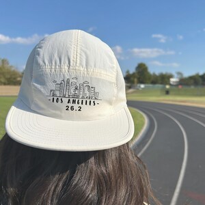 May include: White running hat with a black Los Angeles skyline graphic and the text "Los Angeles - 26.2".