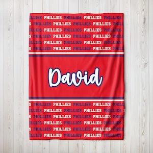 May include: Red blanket with the name "David" in white script. The blanket features the word "PHILLIES" in a repeating pattern in navy blue and white, a design associated with the Philadelphia Phillies.