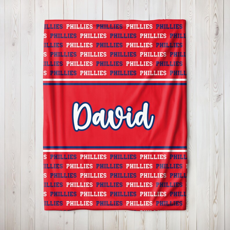 May include: Red blanket with the name "David" in white script. The blanket features the word "PHILLIES" in a repeating pattern in navy blue and white, a design associated with the Philadelphia Phillies.